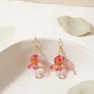 May include: A pair of dangle earrings featuring translucent pink and orange flower-shaped charms with gold-toned wire detailing. Each earring is finished with a small, iridescent spherical bead suspended from the base of the flower and a gold hook.