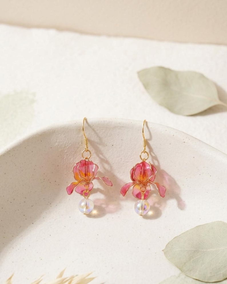 May include: A pair of dangle earrings featuring translucent pink and orange flower-shaped charms with gold-toned wire detailing. Each earring is finished with a small, iridescent spherical bead suspended from the base of the flower and a gold hook.