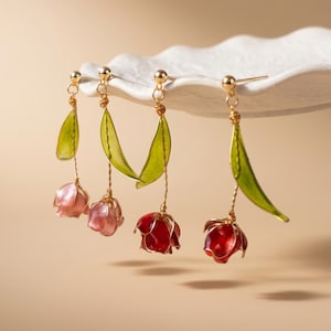 May include: Four drop earrings featuring delicate flower designs suspended from gold-toned metal wires. Two earrings have light pink petals, and two have deep red petals. Each earring includes a single translucent green leaf attached to the stem.