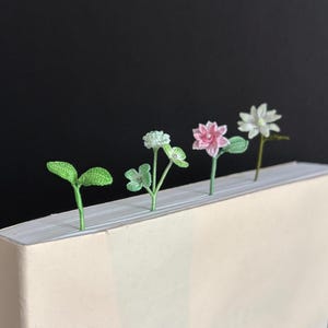 May include: A row of handmade crochet bookmarks featuring various floral designs, including a sprout, clover, pink flower, and white flower. The bookmarks are green, pink, and white, and are placed on top of a book with a light beige cover.
