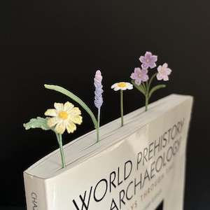 Puede incluir: Un libro con el título "World Prehistory Archaeology" con cuatro marcadores de libros de flores de ganchillo que sobresalen de la parte superior. Los marcadores son una flor de lavanda, una margarita, una flor morada y una margarita amarilla.