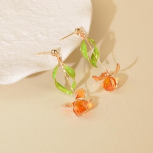 May include: A pair of gold-tone drop earrings featuring a delicate green enameled leaf design attached to a thin metal stem, ending in a translucent orange flower-shaped pendant. The earrings include a standard post back for secure attachment.