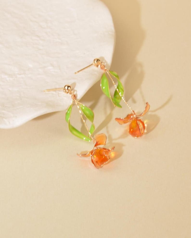 May include: A pair of gold-tone drop earrings featuring a delicate green enameled leaf design attached to a thin metal stem, ending in a translucent orange flower-shaped pendant. The earrings include a standard post back for secure attachment.