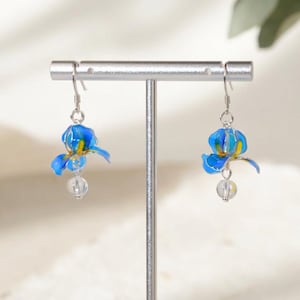 May include: A pair of drop earrings featuring blue and yellow iris flower charms suspended from silver-toned hooks, with a small, clear, spherical bead dangling beneath each flower, displayed on a silver-toned T-shaped jewelry stand.