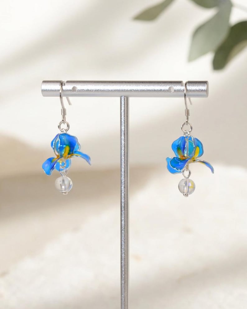 May include: A pair of drop earrings featuring blue and yellow iris flower charms suspended from silver-toned hooks, with a small, clear, spherical bead dangling beneath each flower, displayed on a silver-toned T-shaped jewelry stand.