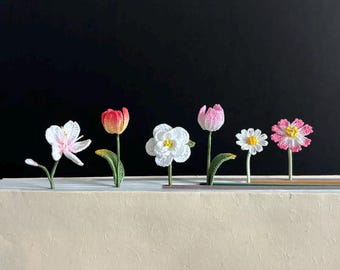 Puede incluir: Cinco marcapáginas de ganchillo en forma de flores de diferentes colores y estilos. Los marcapáginas son rosas, blancos y rojos. Están dispuestos en una fila sobre un libro.