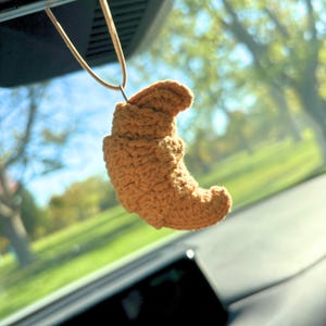 May include: A crocheted, tan-colored croissant car charm hanging from a tan cord. The charm is suspended in front of a blurred background of trees and a bright blue sky, suggesting an outdoor setting.