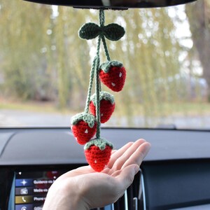 May include: A handmade car charm featuring four crocheted strawberries. The strawberries are red with white seed details and green tops. The charm is suspended from a green crocheted bow and string, designed to hang from a rearview mirror.