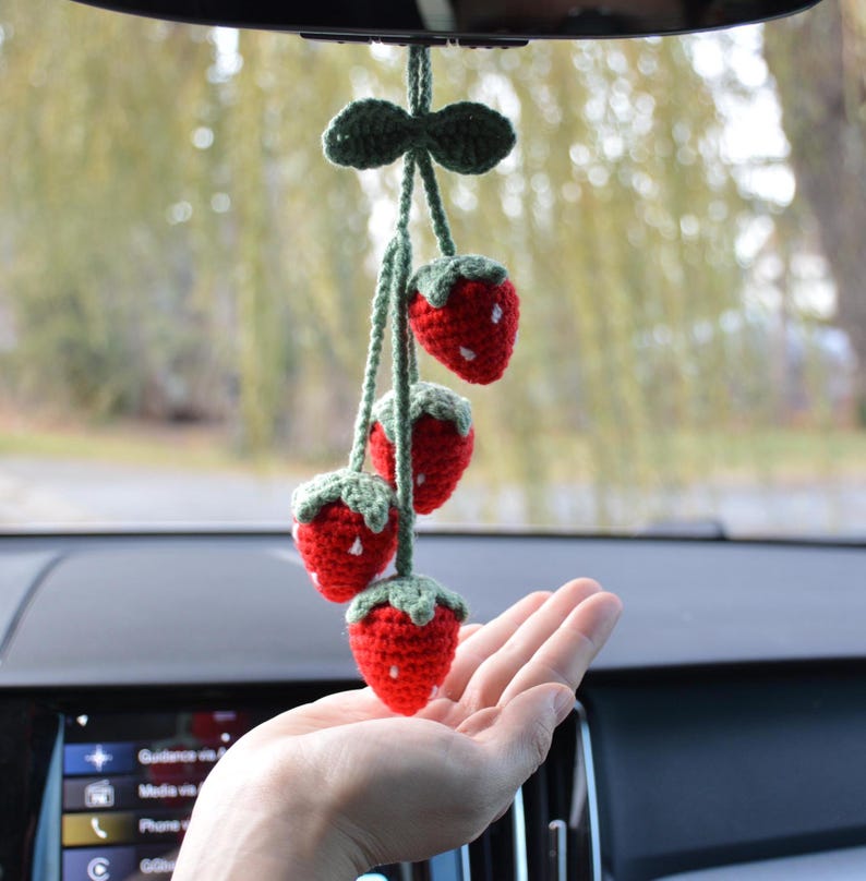 May include: A handmade car charm featuring four crocheted strawberries. The strawberries are red with white seed details and green tops. The charm is suspended from a green crocheted bow and string, designed to hang from a rearview mirror.