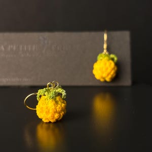 May include: A pair of handmade crochet pineapple earrings. The earrings feature a bright yellow pineapple with a green leafy top. The earrings are suspended from gold-colored hoops. The background is dark, highlighting the vibrant colors of the earrings.