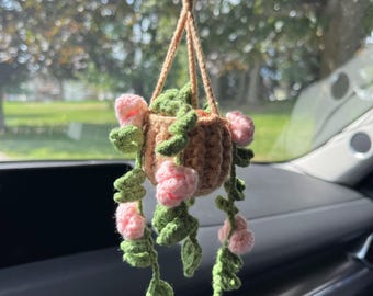 May include: A crocheted hanging basket with pink flowers and green leaves. The basket is brown and suspended by a tan string. The flowers are a soft pink, and the leaves are a vibrant green. This handmade item is a decorative accent.