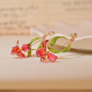 May include: A pair of drop earrings featuring delicate, translucent pink and red flower petals attached to thin, curved green stems. The floral elements are crafted with a metallic wire frame and small gold-toned beads at the top of each earring.
