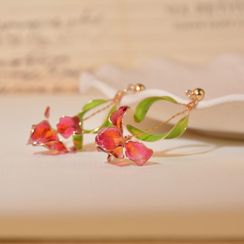 May include: A pair of drop earrings featuring delicate, translucent pink and red flower petals attached to thin, curved green stems. The floral elements are crafted with a metallic wire frame and small gold-toned beads at the top of each earring.