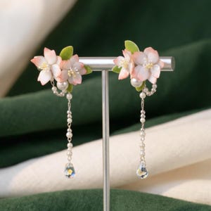 Può includere: Un paio di orecchini pendenti con fiori di ciliegio rosa pallido e foglie verdi, arricchiti da perle bianche e una catenella color argento. Ogni orecchino termina con una perla di cristallo trasparente a goccia sospesa alla catena.