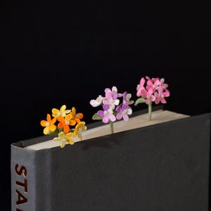 Op de afbeelding: Drie gehaakte bloemenbladwijzers in een donkergrijs boek. De eerste is geel en oranje, de tweede paars en wit, en de derde roze. Elke bloementros is voorzien van kleine witte kraaltjes in het midden en een dunne groene steel.