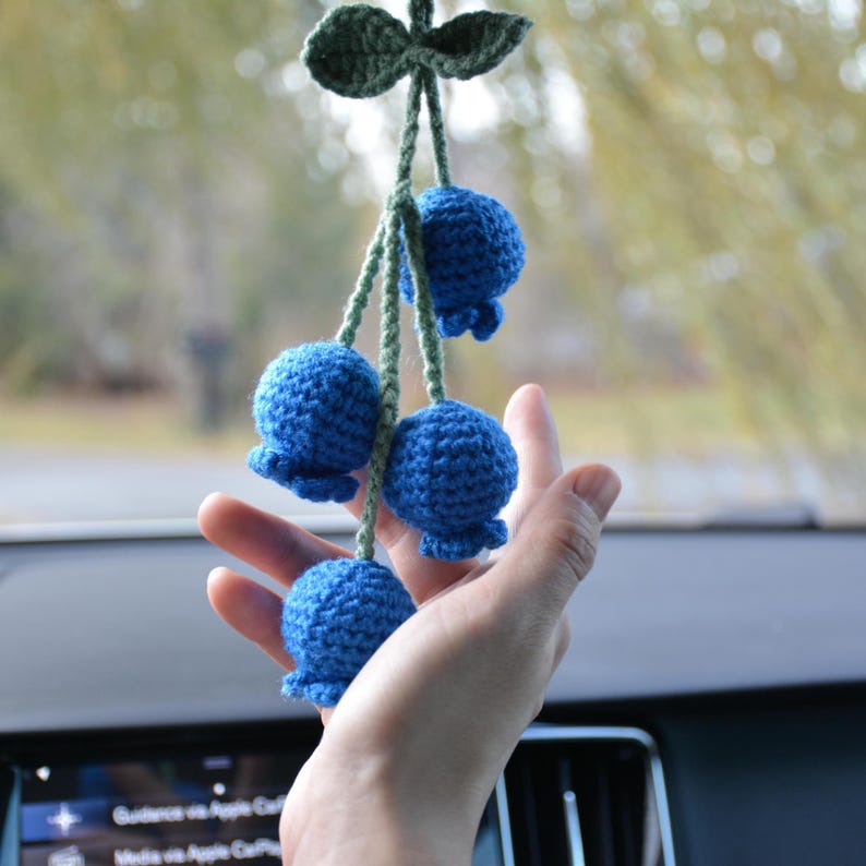 May include: A handmade crochet blueberry car charm. The charm features four blue crocheted blueberries hanging from green stems, with a green leaf at the top. The charm is designed to hang from a car's rearview mirror.