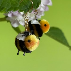 Puede incluir: Un par de pendientes de abeja hechos a mano. Cada pendiente presenta una abeja esponjosa, amarilla y negra con pequeñas patas negras y un ala blanca. Los pendientes cuelgan de ganchos dorados, con un fondo verde.
