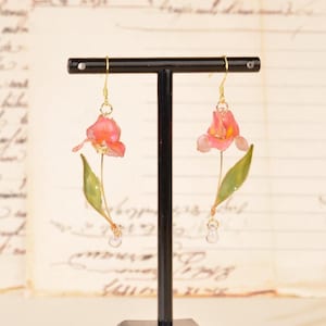 May include: A pair of floral drop earrings. Each earring features a pink and yellow flower with a green leaf and a clear, teardrop-shaped bead. The earrings are displayed on a black stand against a vintage background.