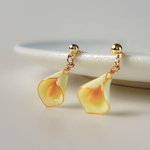 May include: A pair of gold dangle earrings featuring a delicate white and yellow flower design. The earrings are made with a gold chain and a small gold ball at the top.
