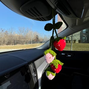 May include: A handmade crochet car charm featuring three strawberries. The strawberries are red and pink with green leaves and stems. The charm hangs from a green crocheted string, adding a touch of whimsy to the car's interior.