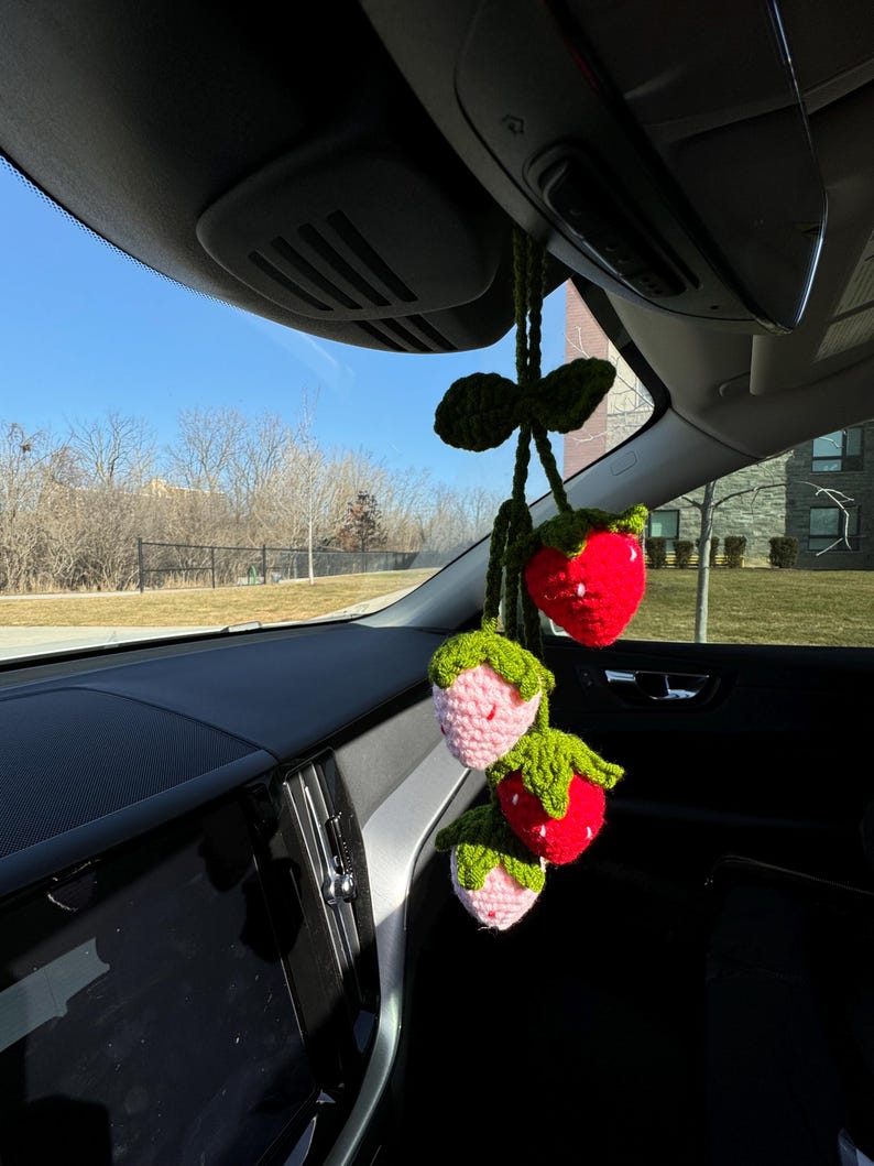 May include: A handmade crochet car charm featuring three strawberries. The strawberries are red and pink with green leaves and stems. The charm hangs from a green crocheted string, adding a touch of whimsy to the car's interior.