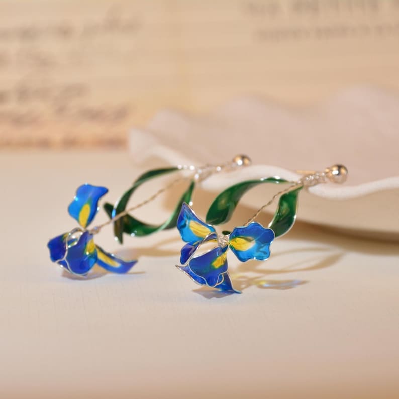 May include: A pair of drop earrings featuring translucent blue and yellow iris flower petals with green leaves, crafted with thin silver-toned wire frames and small spherical stud posts, capturing the delicate form of a blooming iris flower.