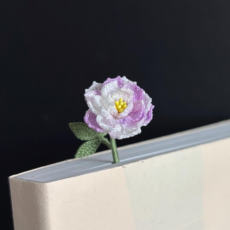 May include: A crocheted flower bookmark with a white and purple flower and green leaves. The bookmark is placed in a book.
