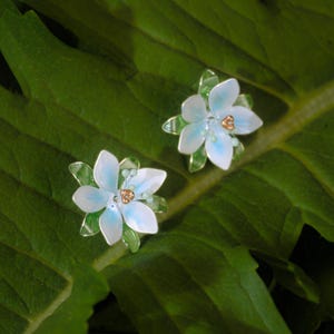 Puede incluir: Un par de pendientes de botón con forma de flores de seis pétalos con pétalos en degradado blanco y azul claro, acentos translúcidos en forma de hoja verde en la base y un pequeño grupo de cuentas metálicas doradas en el centro.