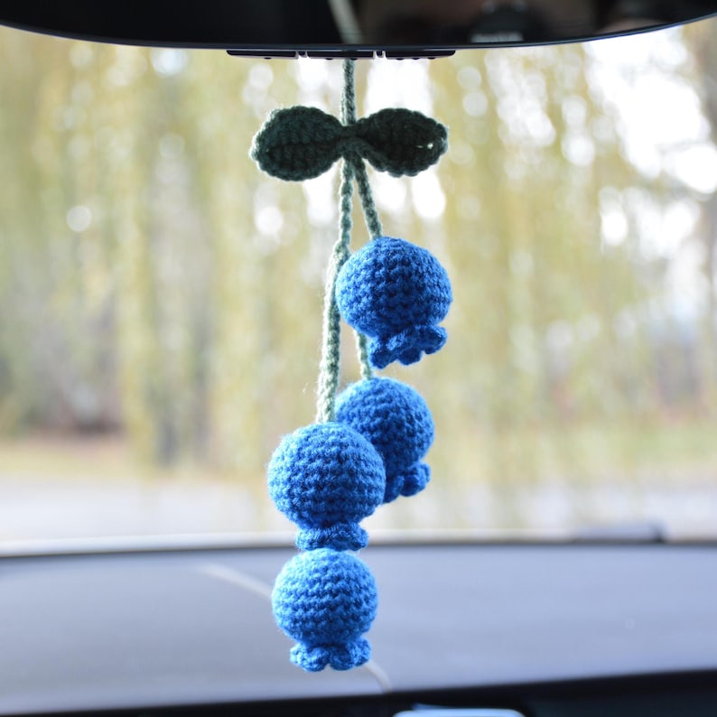 May include: A handmade car charm featuring a cluster of blue crochet berries hanging from a green stem and leaf. The berries are round with a ruffled edge, and the charm is suspended from a car's rearview mirror.