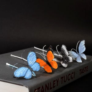 May include: Four decorative butterfly ornaments on thin white stems, featuring textured wings in bright blue, vibrant orange, black and white, and pale blue, arranged in a row on a gray book titled STANLEY TUCCI TASTE.