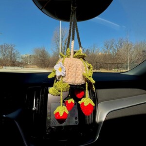 May include: A handmade crochet car charm featuring a beige pot with green leaves and a white flower. Hanging below are three red crochet strawberries with green tops. The charm is suspended from the rearview mirror.