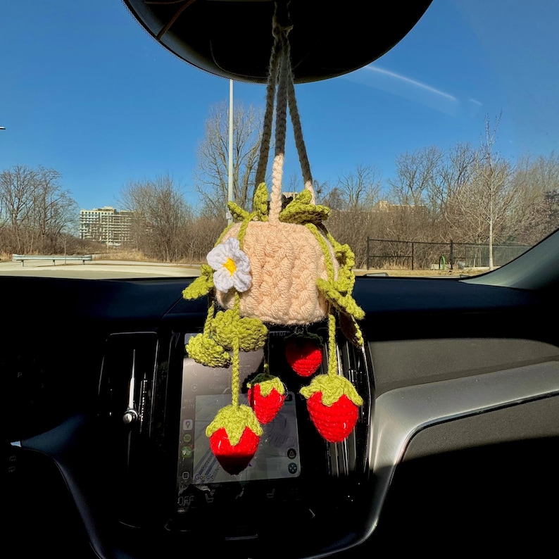 May include: A handmade crochet car charm featuring a beige pot with green leaves and a white flower. Hanging below are three red crochet strawberries with green tops. The charm is suspended from the rearview mirror.