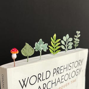 Può includere: Un libro intitolato "World Prehistory Archaeology" con un fungo rosso e bianco, una foglia di monstera verde, un cespuglio verde, una felce verde, un cespuglio verde e una foglia di eucalipto verde come segnalibro.