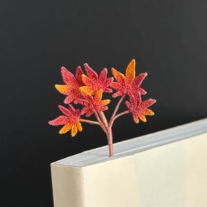 Könnte beinhalten: Ein gehäkeltes Lesezeichen in Form eines kleinen Baumes mit orangefarbenen und roten Blättern. Das Lesezeichen befindet sich oben auf einem weißen Buch.
