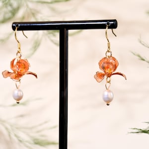May include: A pair of gold-toned hook earrings featuring translucent orange flower-shaped charms and dangling white pearls. The floral elements have delicate, layered petals with a glossy finish, suspended from thin gold wires and metal hooks.