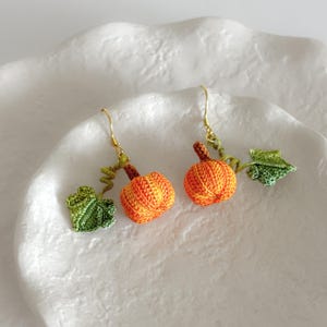 May include: A pair of crocheted pumpkin earrings with green leaves. The earrings are made with orange, yellow, and green yarn.