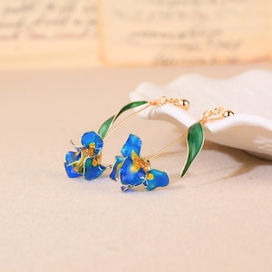 May include: A pair of floral earrings featuring blue and yellow flower pendants with gold-colored accents. The earrings have green leaf details and gold-colored ear wires. The earrings are designed to resemble a flower.