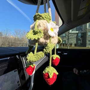 May include: A handmade crochet car charm featuring a beige pot with green leaves and a white flower. Three red crochet strawberries hang below. The charm is suspended from the car's rearview mirror.