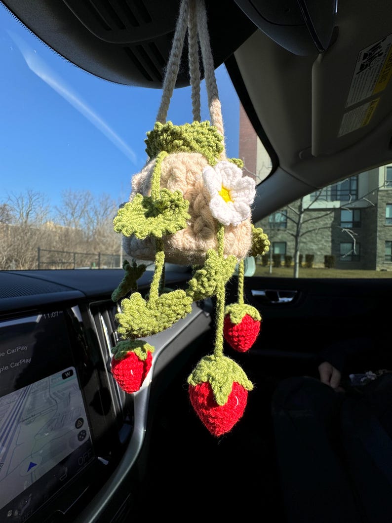 May include: A handmade crochet car charm featuring a beige pot with green leaves and a white flower. Three red crochet strawberries hang below. The charm is suspended from the car's rearview mirror.