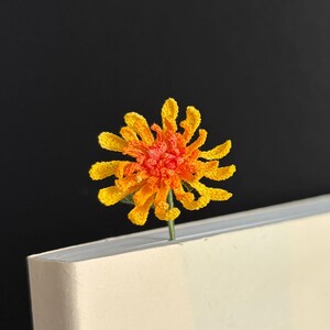 May include: A crocheted orange and yellow flower bookmark with a green stem.