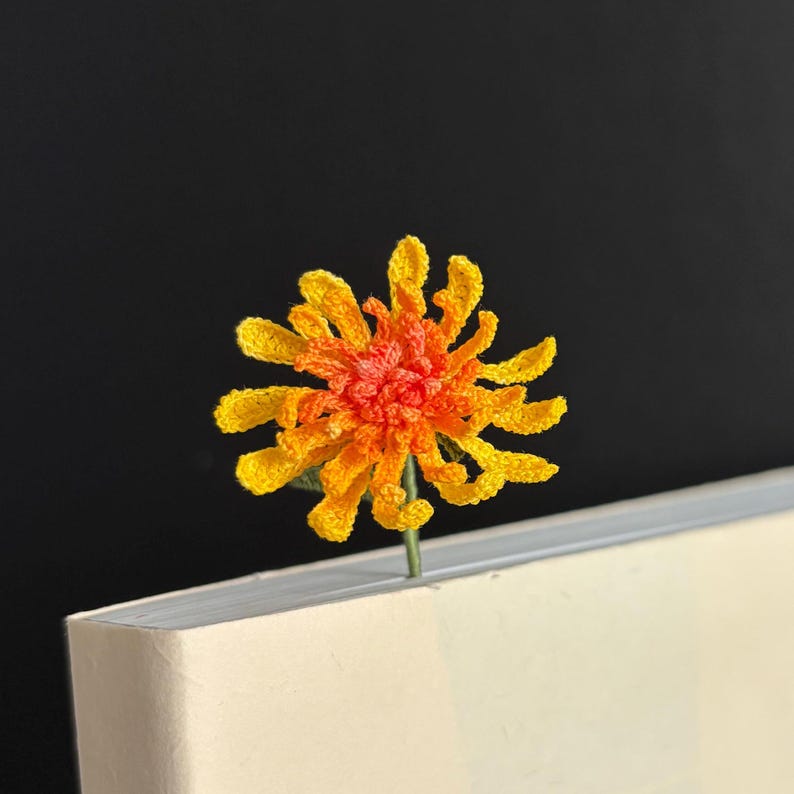 May include: A crocheted orange and yellow flower bookmark with a green stem.