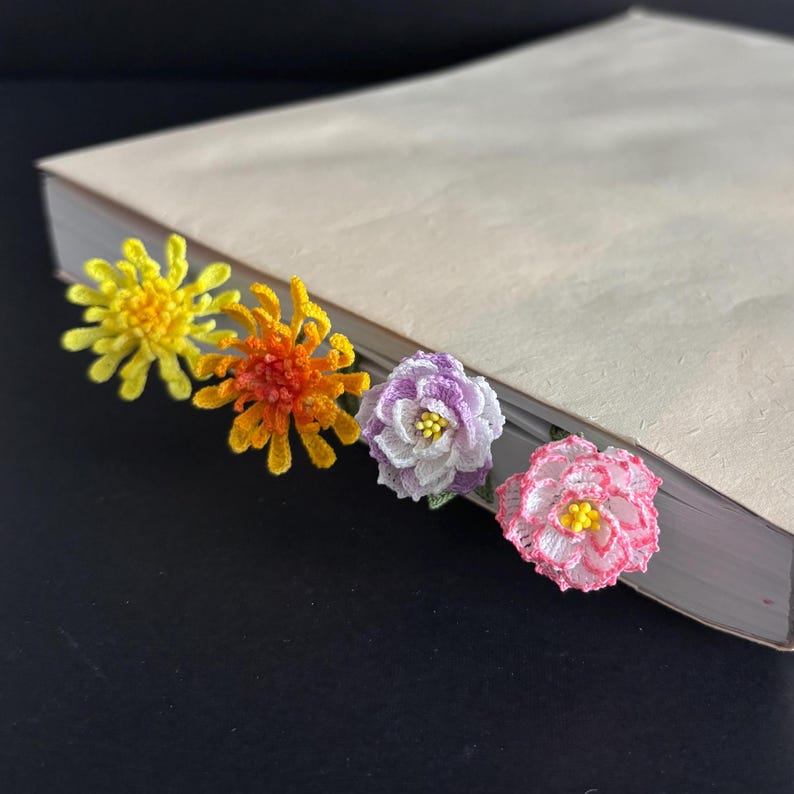 May include: Four crocheted flower bookmarks in different colors: yellow, orange, purple, and pink. The bookmarks are attached to a white book.