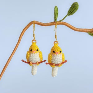 May include: A pair of handmade crochet bird earrings. Each earring features a yellow and white bird with a small red beak, perched on a brown branch. The earrings have gold-colored hooks and are set against a light blue background.