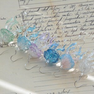 以下が含まれることがあります： 青、紫、緑の色合いの花柄が特徴の手作りのイヤリングのコレクション。各イヤリングは、銀色のフックと、詳細な半透明の花びらを特徴としています。手書きのスクリプトが書かれたヴィンテージ紙の背景に表示されています。