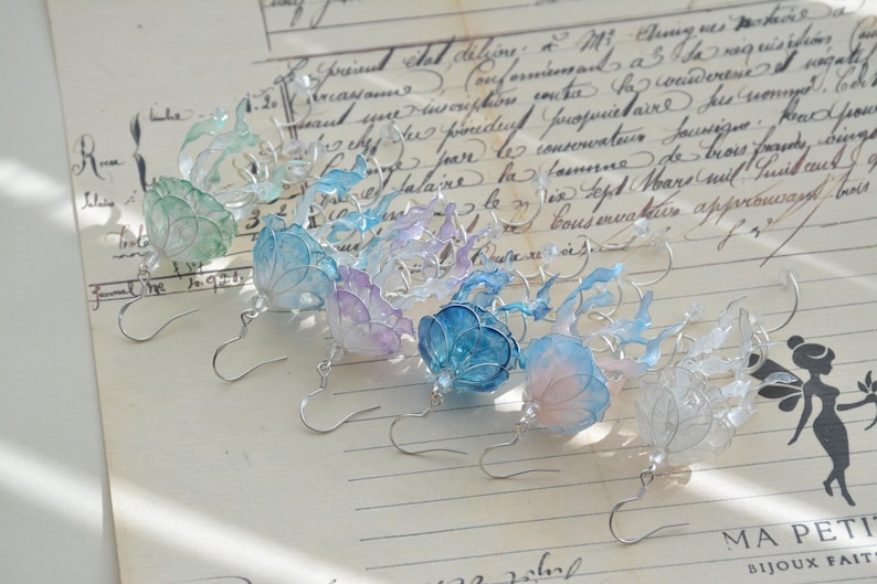 以下が含まれることがあります： 青、紫、緑の色合いの花柄が特徴の手作りのイヤリングのコレクション。各イヤリングは、銀色のフックと、詳細な半透明の花びらを特徴としています。手書きのスクリプトが書かれたヴィンテージ紙の背景に表示されています。