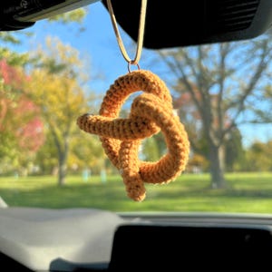 May include: A crocheted pretzel-shaped car charm hanging from a rearview mirror. The charm is a light brown color with a slightly textured surface.