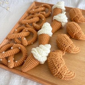 Könnte beinhalten: Eine Sammlung gehäkelter Lebensmittel, darunter Brezeln, Croissants und Eiswaffeln, die auf einem Holztablett präsentiert werden. Die Artikel sind alle in einer hellbraunen Farbe mit weißen Akzenten gehalten.