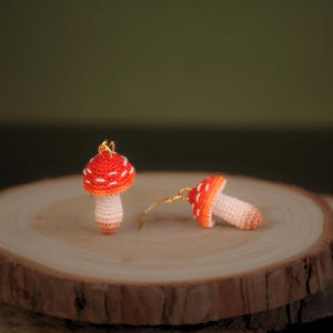 May include: A pair of crocheted mushroom earrings with red and white caps and cream-colored stems. The earrings are hanging from gold hooks.