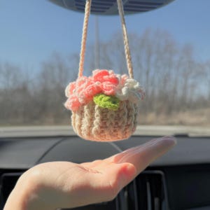 May include: A crocheted flower basket hanging from a rearview mirror. The basket is filled with pink and white flowers and a green leaf.