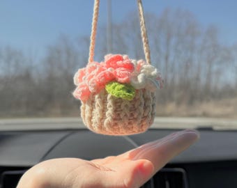 May include: A crocheted flower basket hanging from a rearview mirror. The basket is filled with pink and white flowers and a green leaf. 